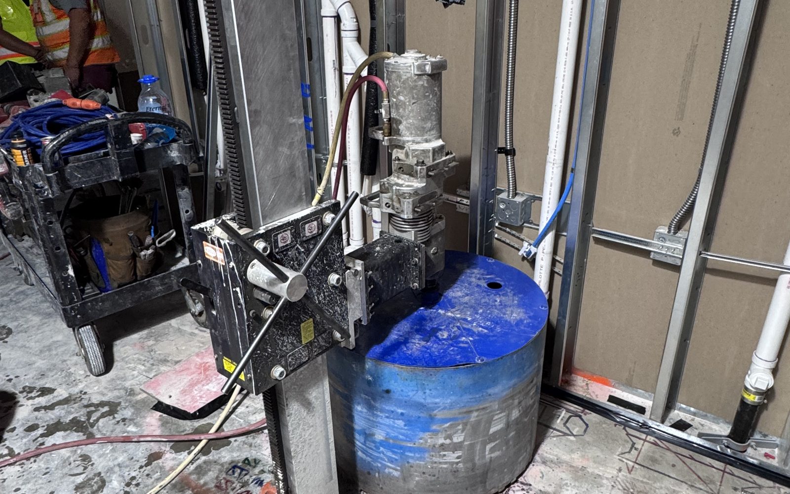 Core drill rig mounted on a stand indoors beside a blue core barrel, metal stud framing behind it
