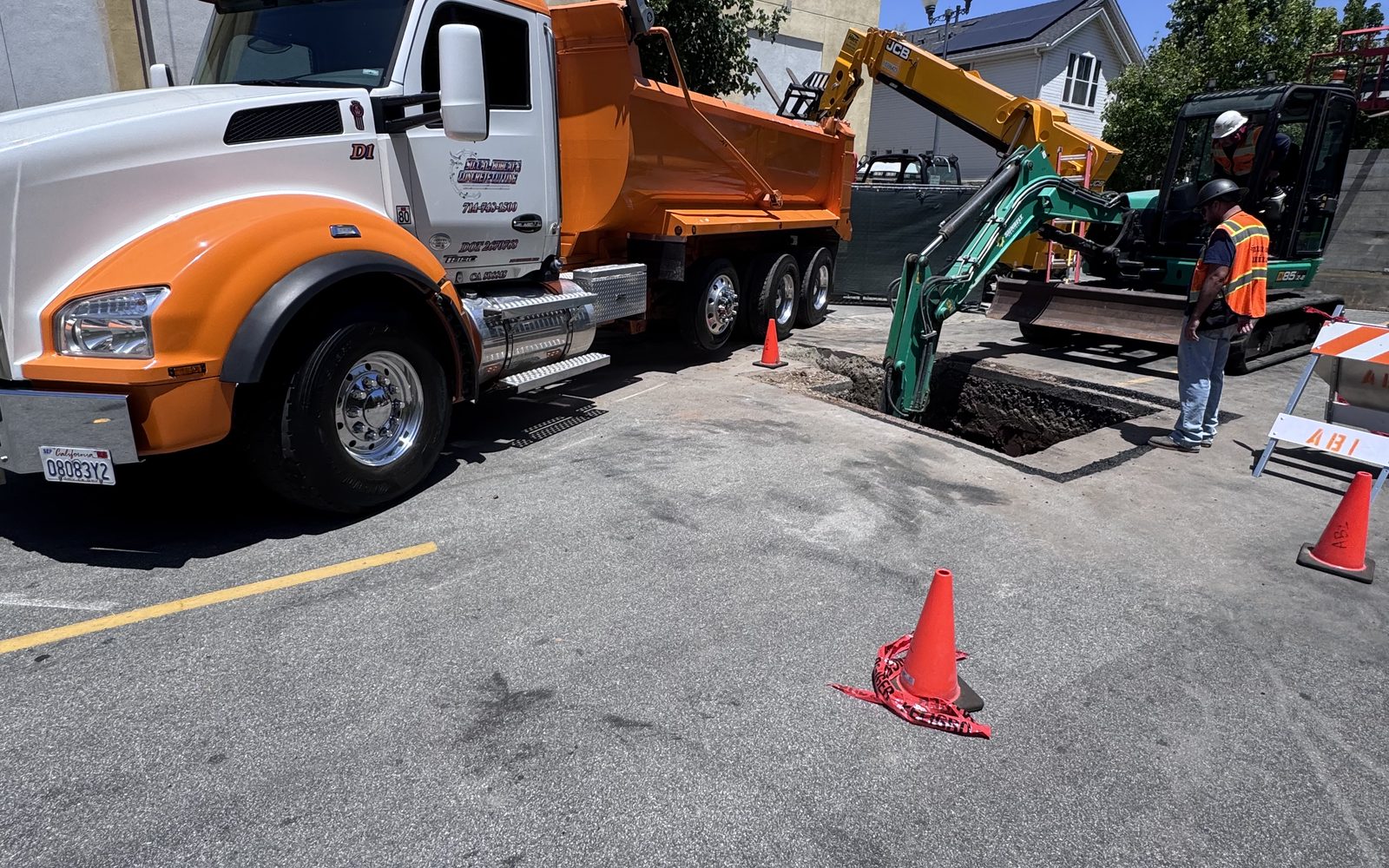 Orange dump truck beside a green mini excavator digging in a parking lot with a worker in a vest