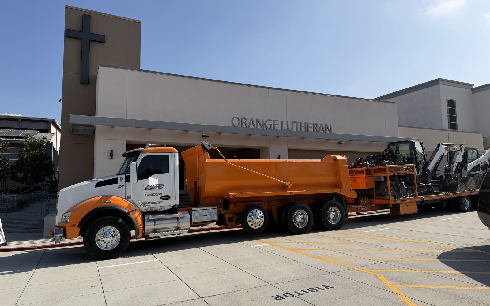 Orange dump truck with two Bobcat skid steers on a trailer parked in front of a building
