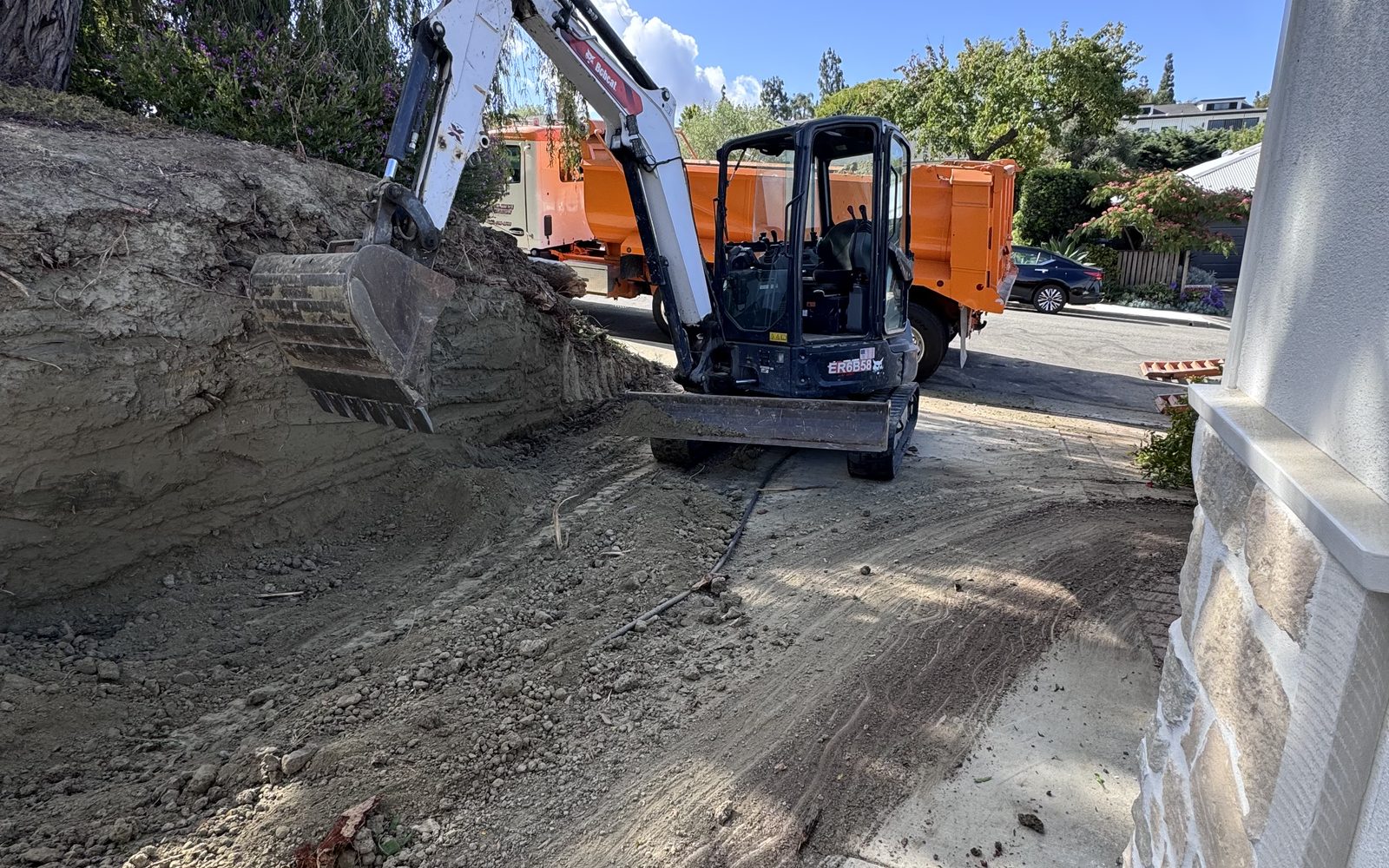 Mini excavator digging beside a house with an orange dump truck parked in the street