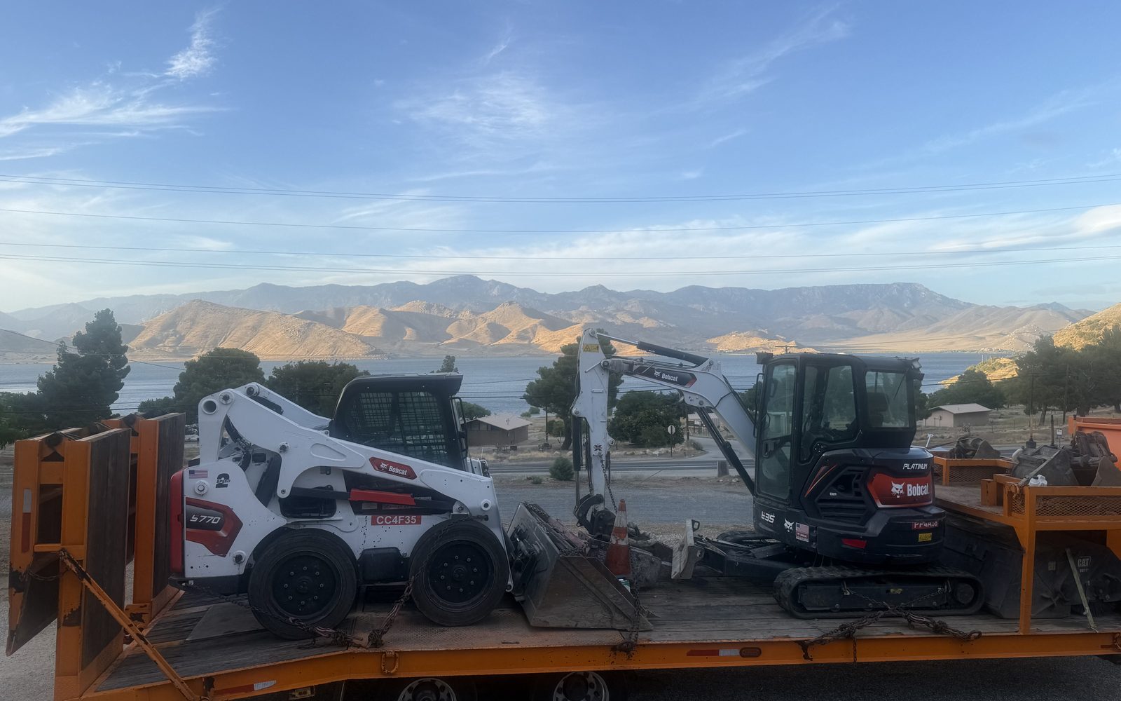 Bobcat skid steer and mini excavator chained down on an orange equipment trailer with a lake and mountains behind