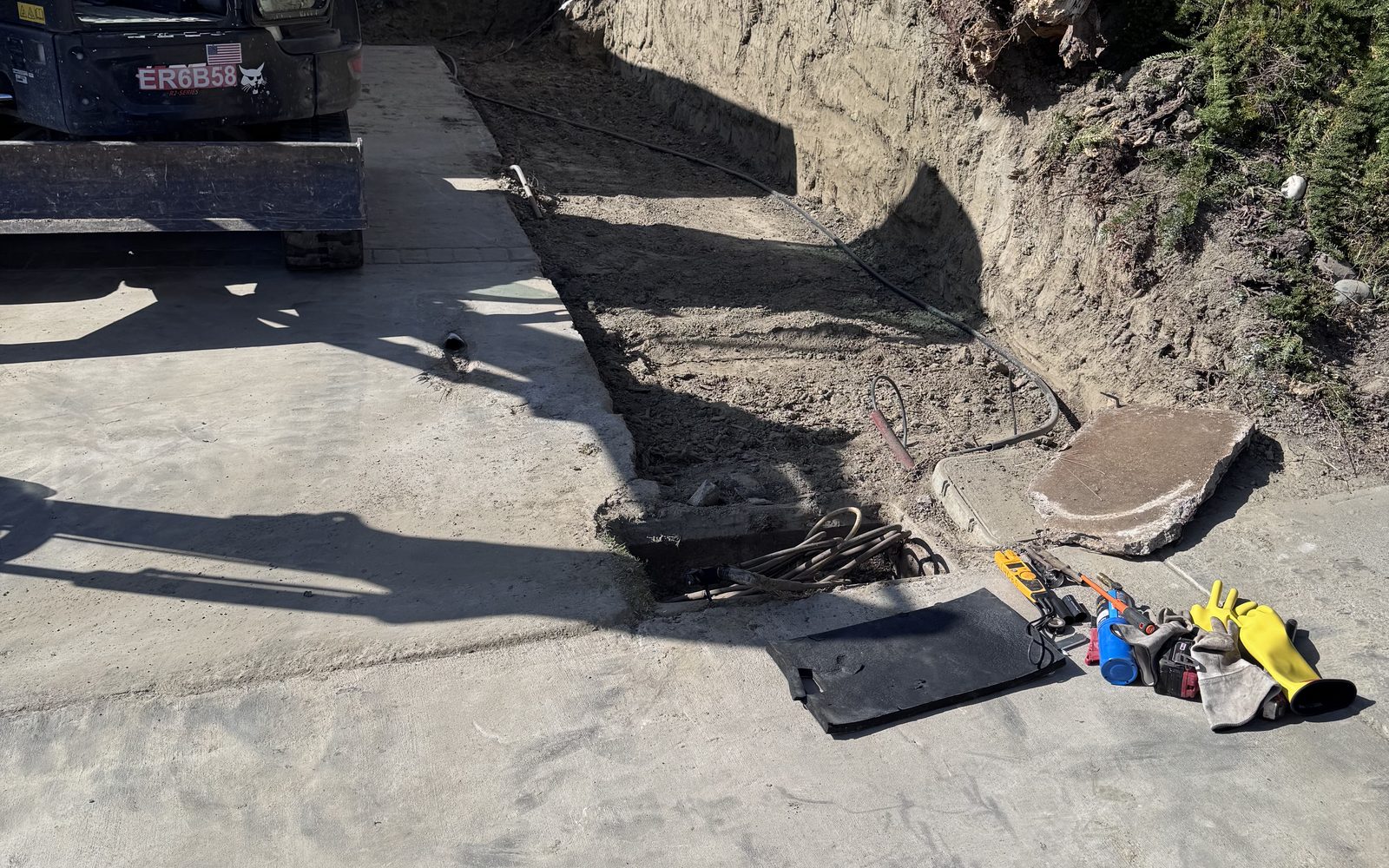 Narrow trench dug alongside a concrete driveway with a mini excavator parked at the end