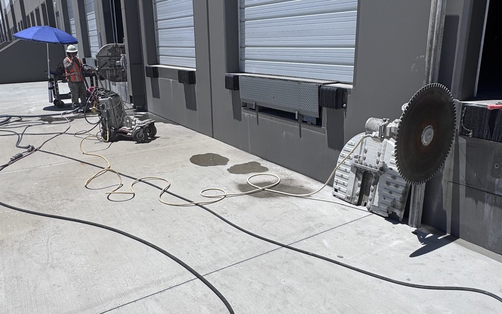 Large wall saw with a diamond blade parked against a loading dock wall, hoses run across the concrete
